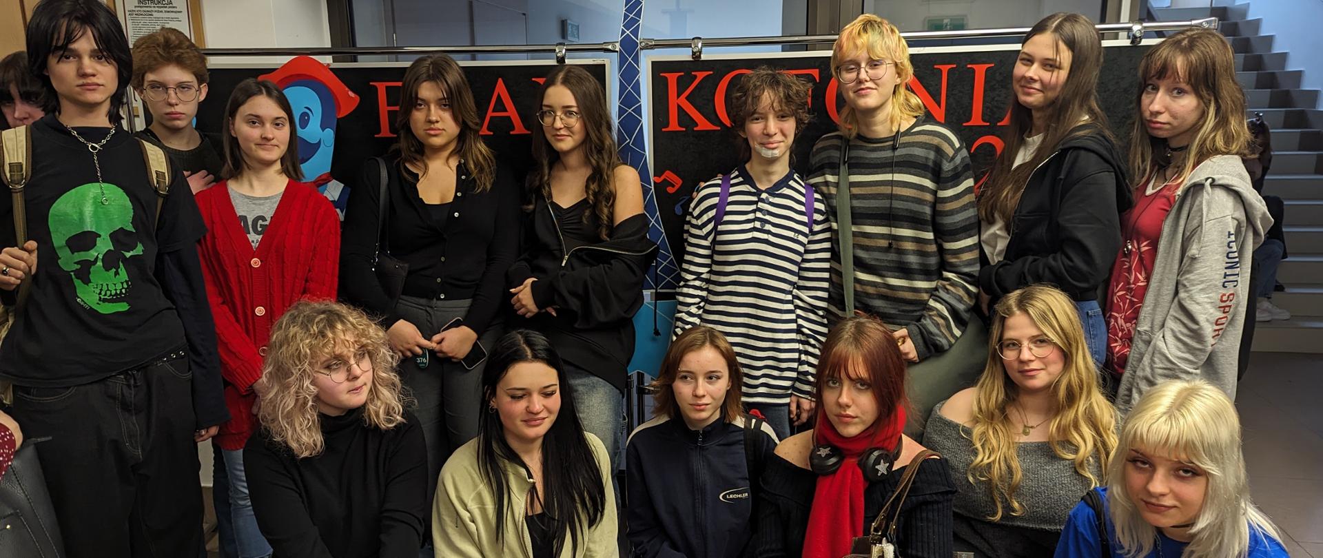 Grupa młodzieży pozuje do fotografii we wnętrzu budynku, w tle schody i szklane drzwi. W pierwszym rzędzie sześć osób kuca lub siedzi, w drugim rzędzie dziesięć osób stoi