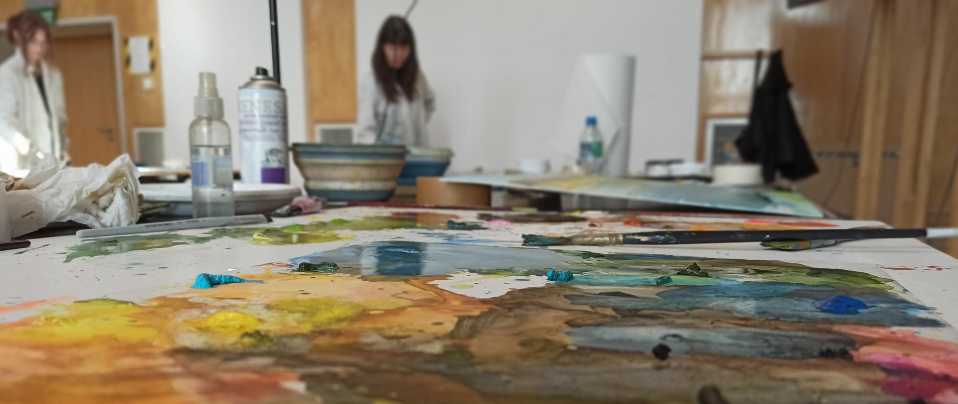 Na zdjęciu widać fragment stołu w pracowni malarskiej pokrytego barwnymi plamami farb wodnych – głównie w odcieniach żółci, błękitu i brązu. Na stole leżą pędzle, tubki i butelka z rozpylaczem. W tle, nieco rozmytym, znajdują się dwie osoby w fartuchach malarskich pracujące nad swoimi obrazami. Całość tworzy atmosferę skupienia i twórczej pracy podczas warsztatów artystycznych.