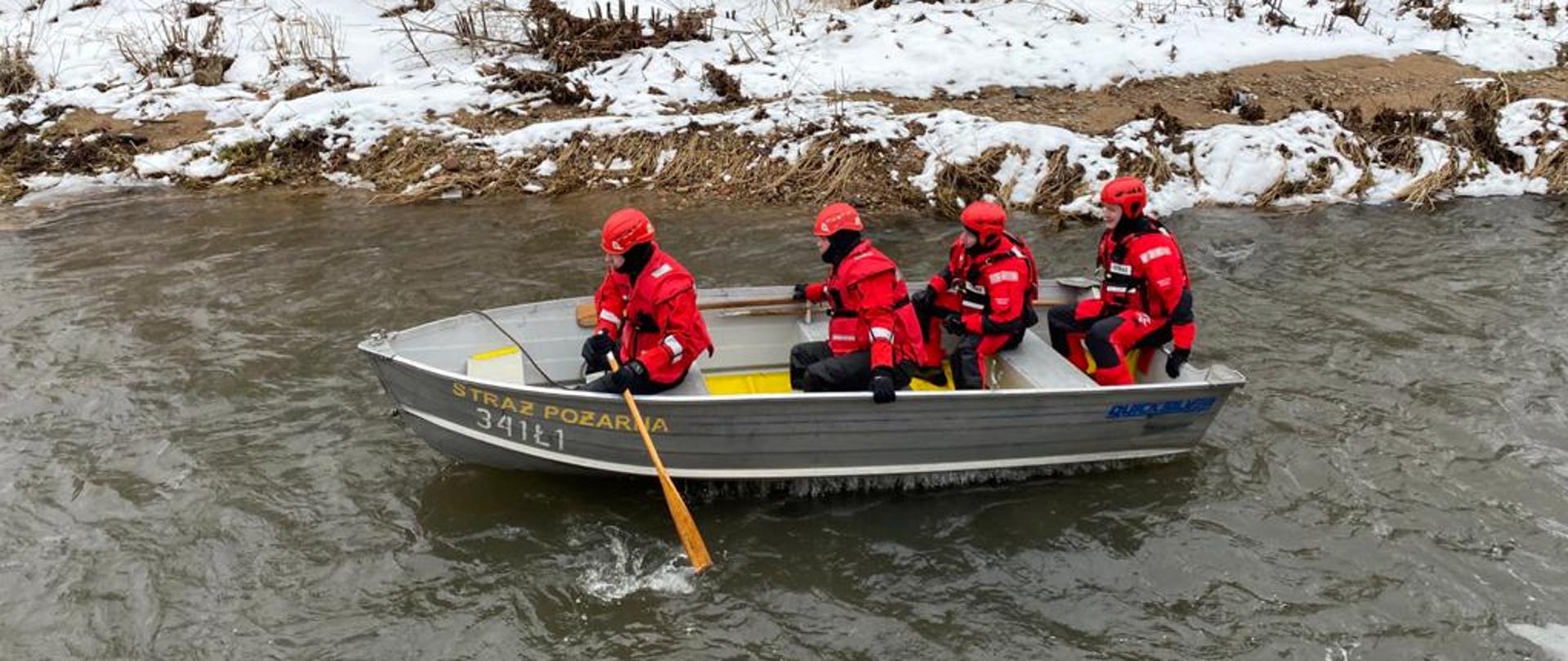 Zdjęcie przedstawia strażaków płynących łódką po rzecze. Strażacy ubrani są w czerwone skafandry oraz czerwone kaski. W łódce znajduje się czterech strażaków. Pierwszy z nich ma wiosło.