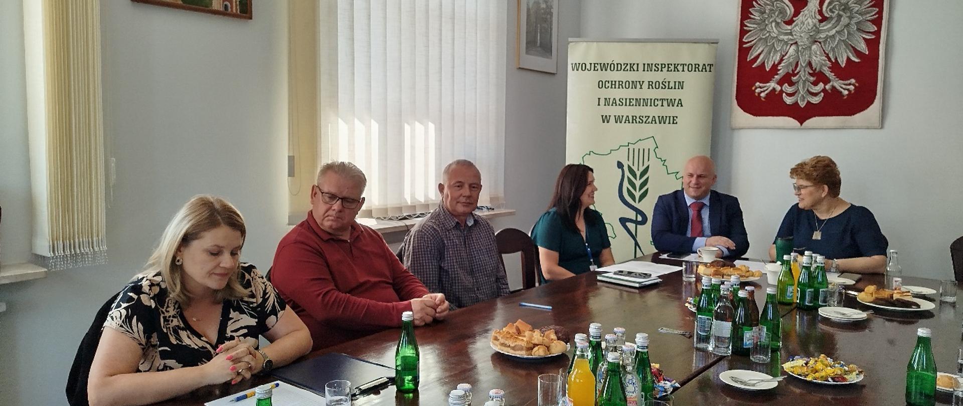 Sześć osób siedzi przy stole konferencyjnym w sali urzędu. Na stole znajdują się dokumenty, napoje oraz poczęstunek. W tle widać baner Wojewódzkiego Inspektoratu Ochrony Roślin i Nasiennictwa w Warszawie oraz godło Polski na ścianie.