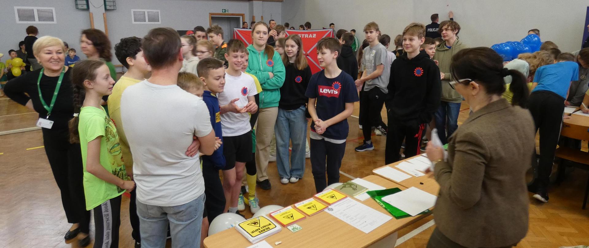 Uczniowie szkoły podstawowej stoją wokół pracownika placówki i stoiska informacyjnego na środku sali gimnastycznej.
