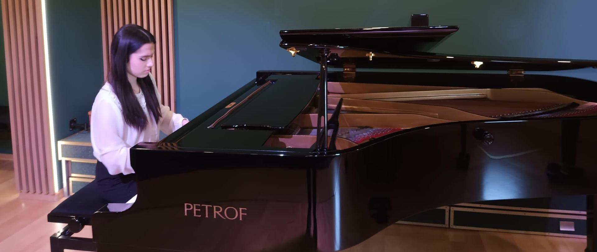 Zdjęcie przedstawia pianistkę grającą na fortepianie za fortepianem widoczna ściana w kolorze zielonym oraz podświetlanymi lamelami