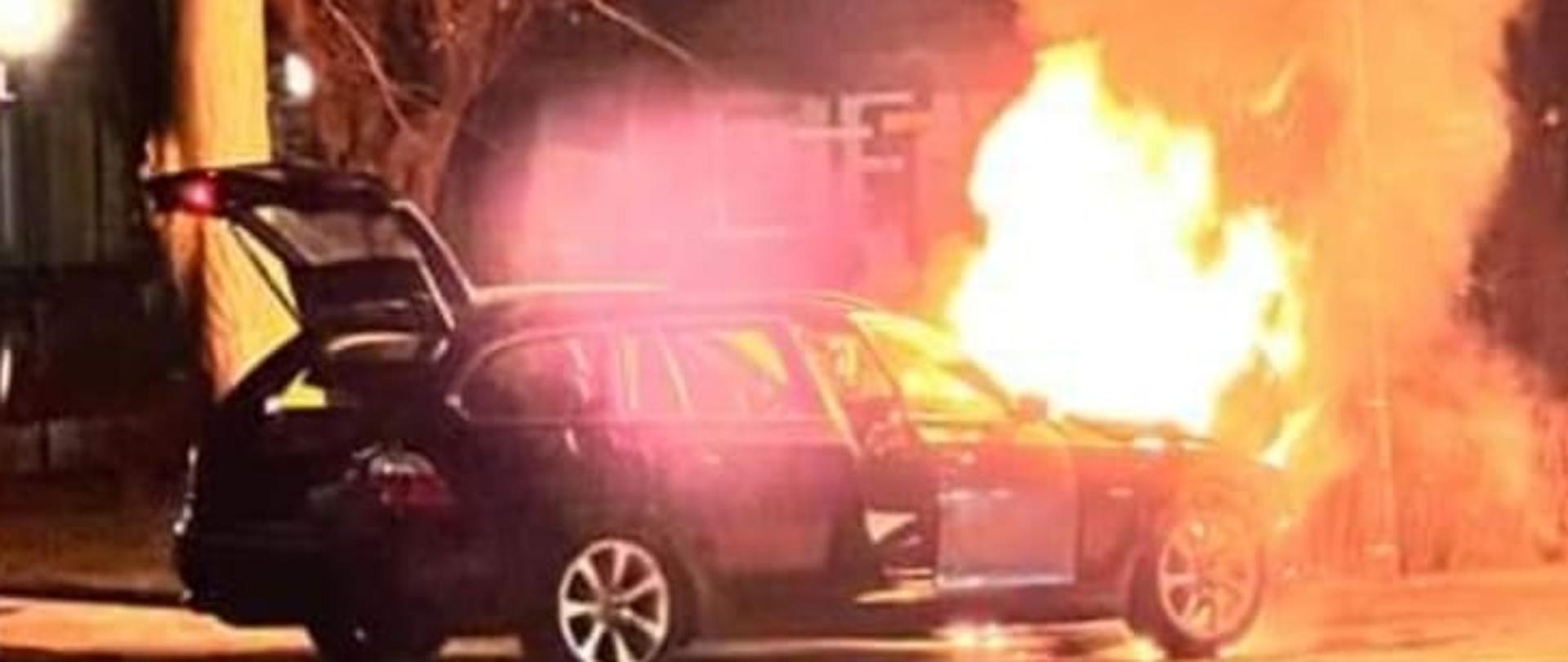 Czarny samochód stoi z otwartymi drzwiami oraz tylną klapę przód pojazdu w ogniu za samochodem widać drzewa