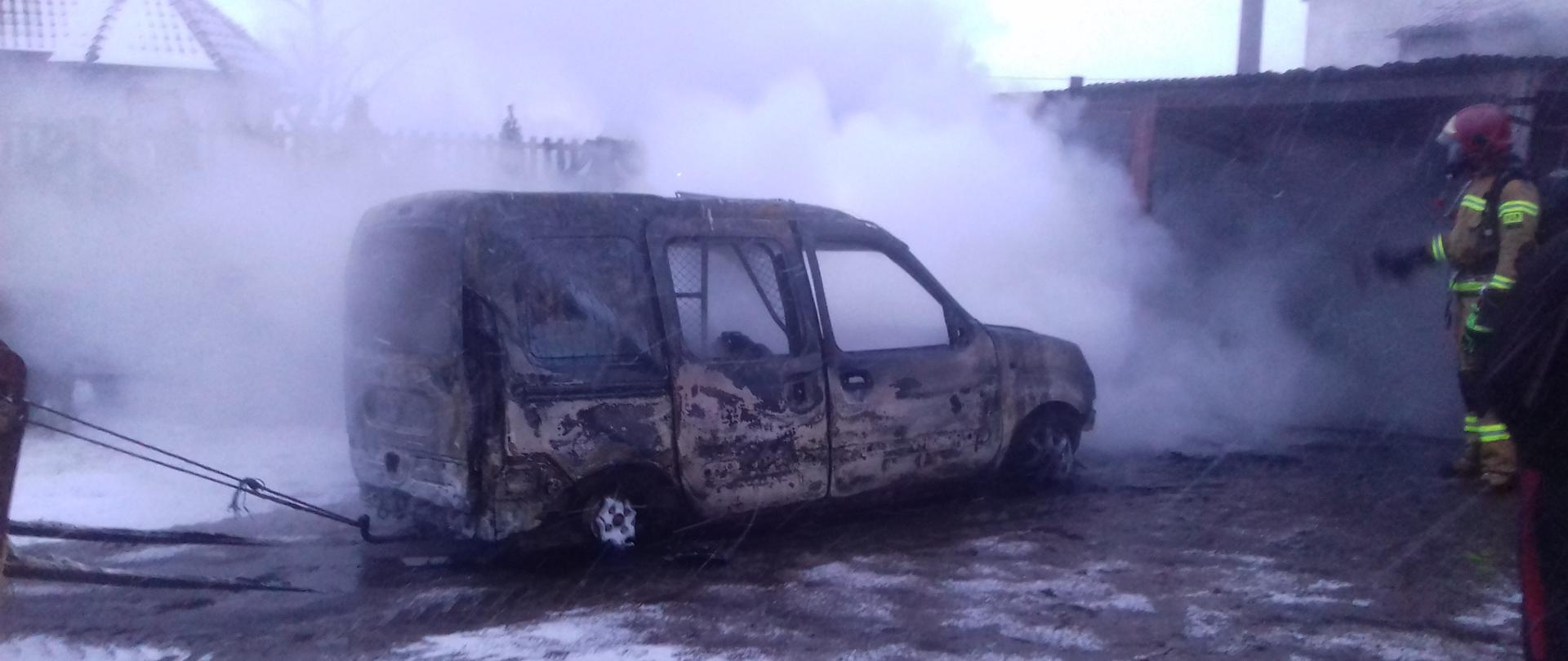 Na zdjęciu widać doszczętnie spalony samochód osobowy, który został wyciągnięty przy użyciu liny na zewnątrz garażu. Za samochodem panuje duże zadymienie. Strażak dogasza zarzewia ognia