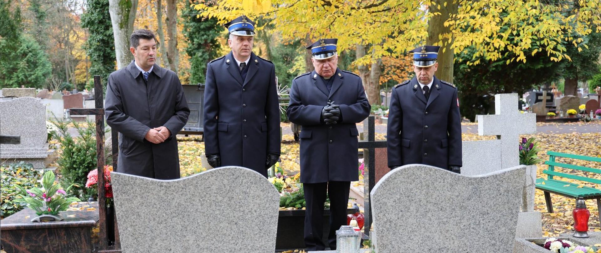 Na zdjęciu widać czterech mężczyzn stojących na cmentarzu przy dwóch sąsiadujących grobach z jasnoszarego granitu. Jest jesień – alejki i płyty nagrobne pokryte są żółtymi liśćmi. Dwóch mężczyzn po prawej stronie ma na sobie granatowe mundury strażackie z granatowymi rogatywkami, podobnie ubrany jest trzeci z lewej. Mężczyzna stojący najbardziej po lewej ma ciemny płaszcz, bez nakrycia głowy. Wszyscy stoją w skupieniu, z powagą, część z nich złożonymi dłońmi, jakby w chwili zadumy lub modlitwy. Na grobach stoją zapalone znicze. W tle widoczne są kolejne nagrobki, drzewa z żółtymi liśćmi oraz zielona ławka po prawej stronie.