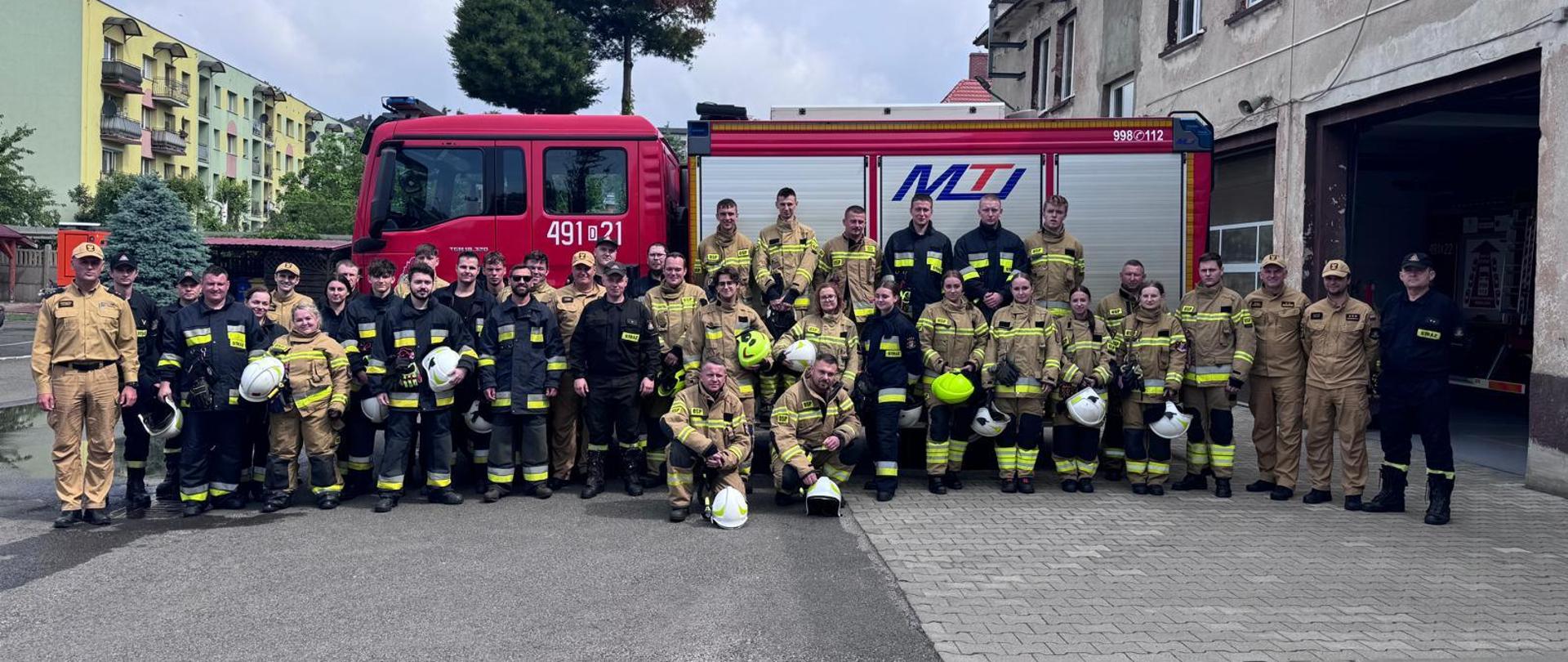 Zdjęcie grupowe po zakończonym egzaminie. Słuchacze oraz egzaminujący stoją na tle samochodu pożarniczego. 
