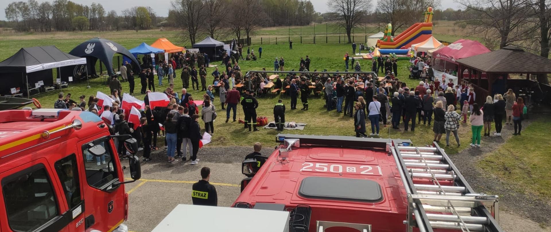 Na zdjęciu plac wypełniony uczestnikami pikniku. Po lewej oraz prawej stronie rozstawione namioty, w głębi po prawej dmuchana zjeżdżalnia. Pośrodku stoliki wraz z ławeczkami
