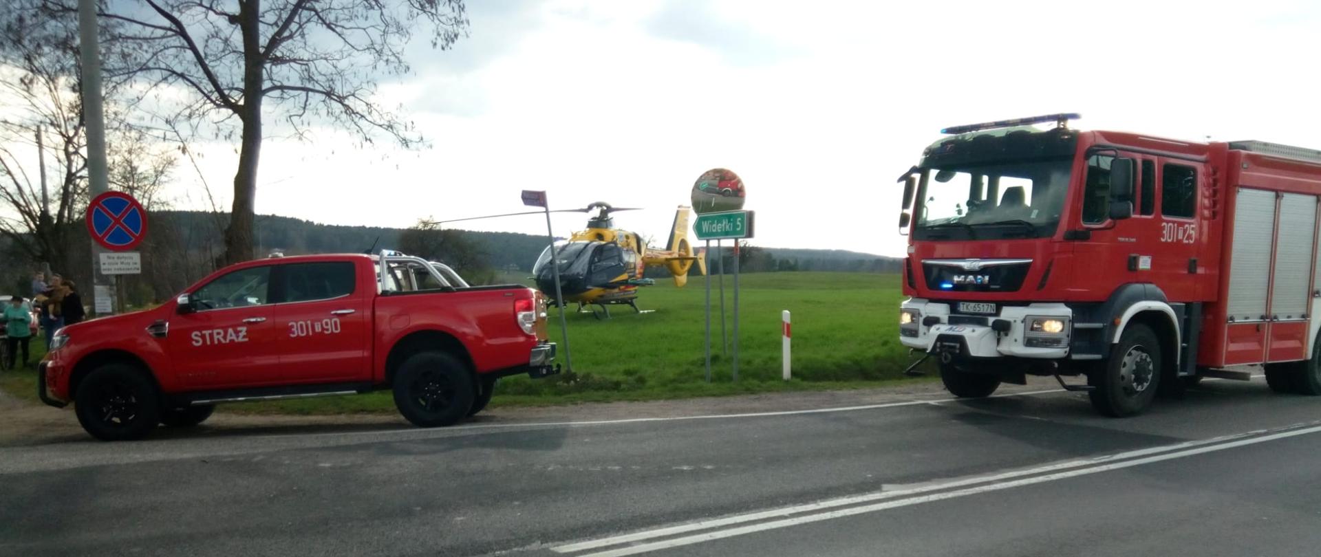 Zdjęcie przedstawia dwa samochody pożarnicze stojące na pasie drogowym drogi krajowej 73. Na pobliskiej łące wylądował śmigłowiec Lotniczego Pogotowia Ratunkowego. 