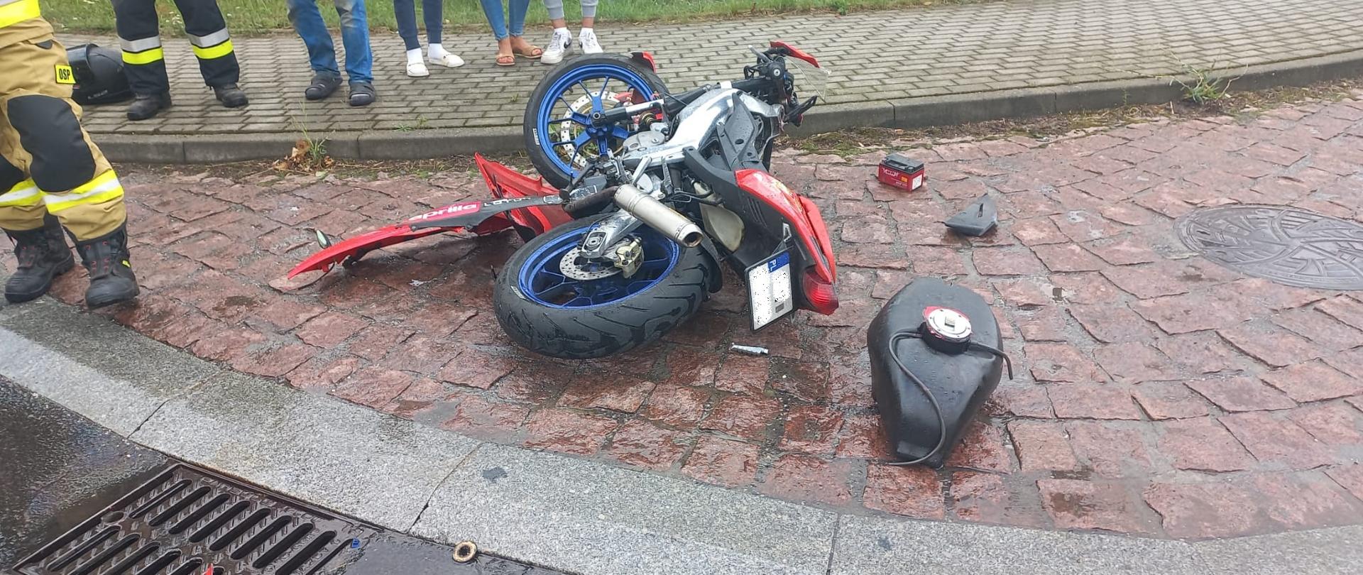 Po środku leżący motocykl po zderzeniu z samochodem osobowym. Na chodniku stoją ludzie,