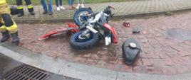 Po środku leżący motocykl po zderzeniu z samochodem osobowym. Na chodniku stoją ludzie,