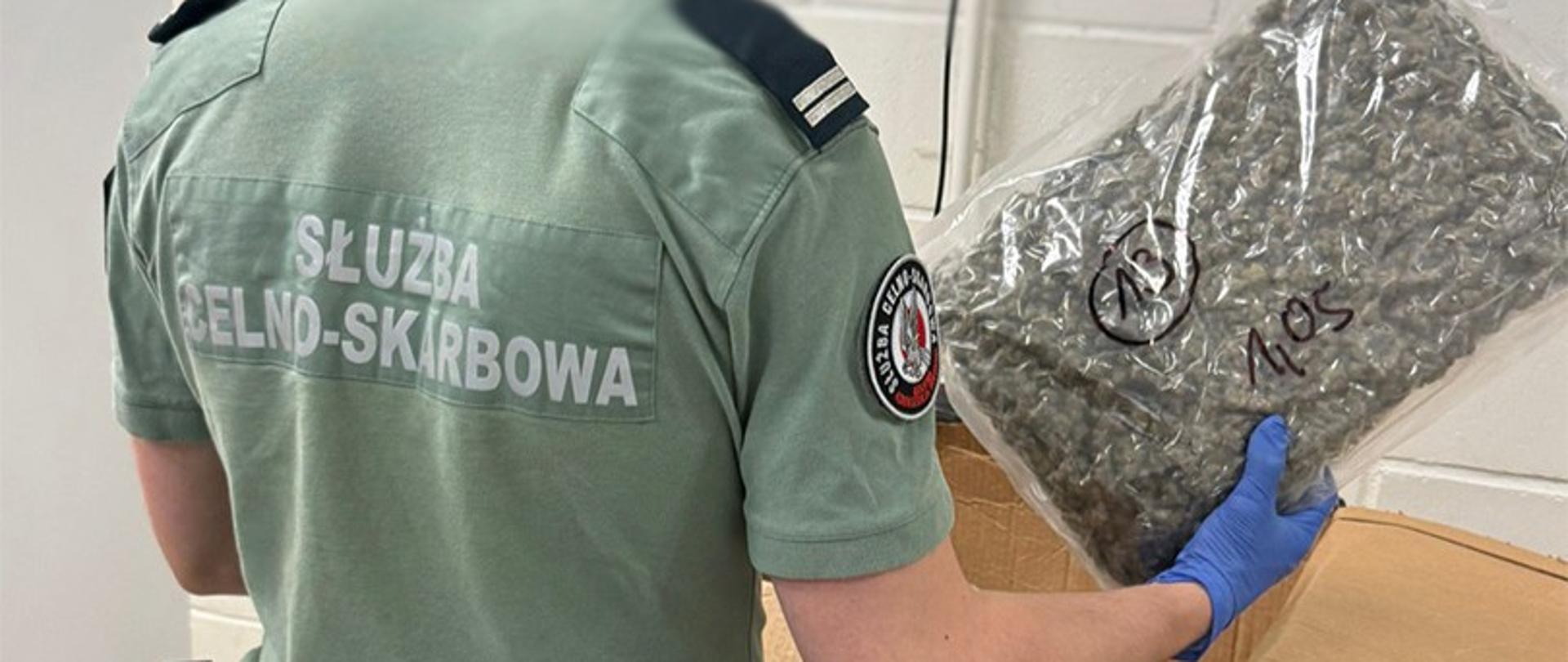 Funkcjonariusz trzyma duży przezroczysty pakunek z rozdrobnioną substancją roślinną; na opakowaniu widoczne są ręczne oznaczenia.