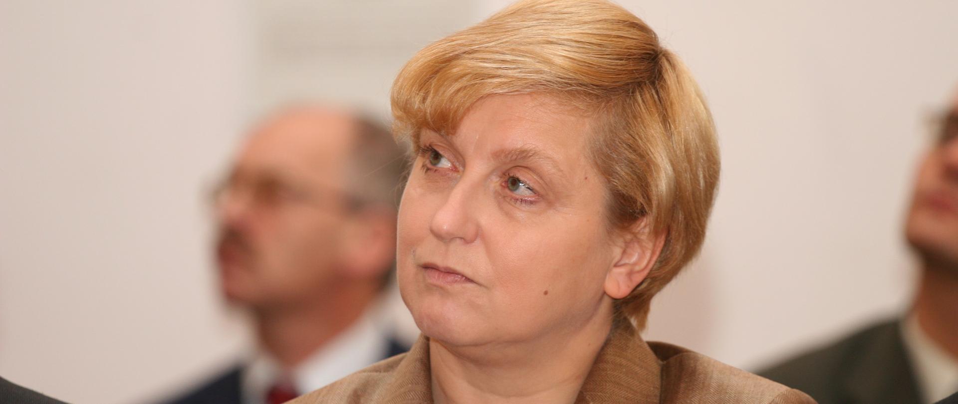 Anna Fotyga kolorowy portret kobiety, krótkie blond włosy, złota marynarka, czarne korale, w tle zamglone postaci mężczyzn.