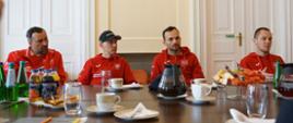 Żużlowcy w czerwonych bluzach sportowych siedzą za stołem. Na stole widoczne są napoje, owoce, woda.