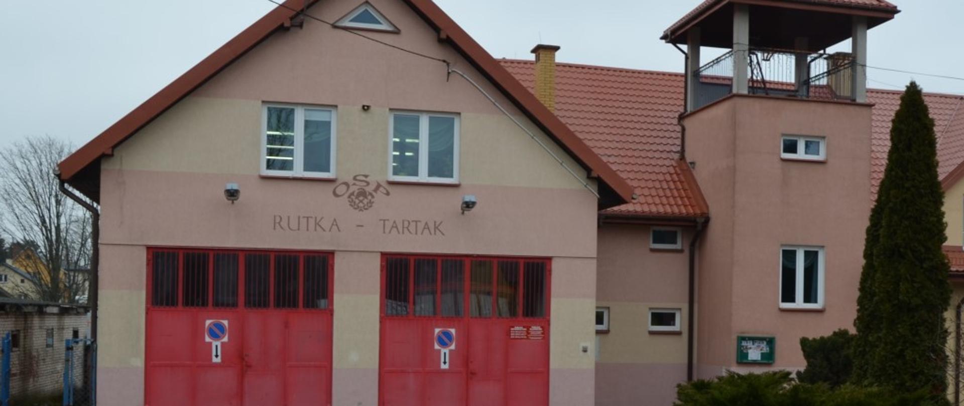 Widoczny budynek w jasno różowych kolorach z dwiema bramami koloru czerwonego. Po prawej stronie widoczna wieża. Dach koloru jasno czerwonego. Podjazd przed budynkiem wyłożony kostką. 