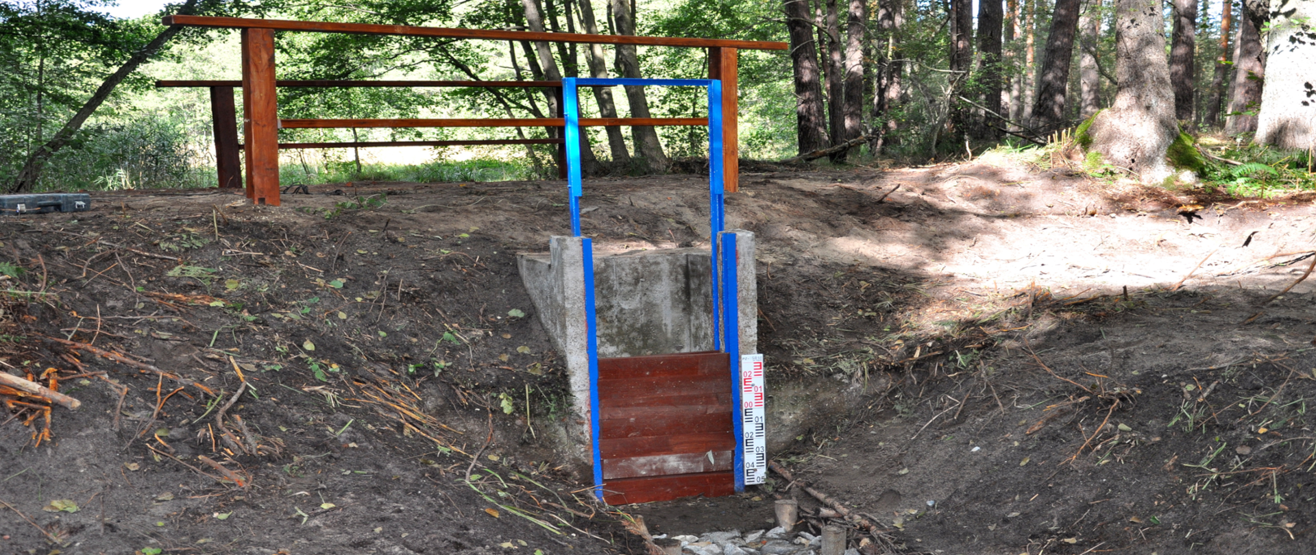 Fragment rowu wypełnionego kamieniami i betonowej konstrukcji. W tle drewniane barierki i las.