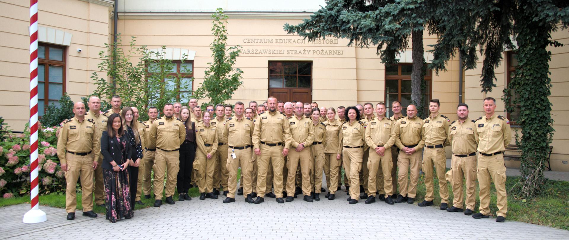 Na zdjęciu wykonano pamiątkową fotografię grupową przed zabytkowym budynkiem. Kilkudziesięciu strażaków stoi w kilku rzędach, ubranych w piaskowe mundury służbowe. W tle widoczne są drzewa, elewacja budynku oraz dziedziniec. Zdjęcie oddaje atmosferę integracji, dumy i wspólnego podsumowania szkolenia.