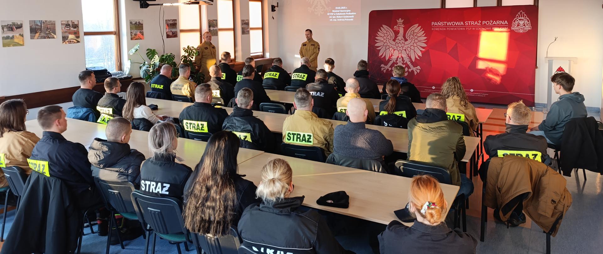 Sala szkoleniowa KP PSP Sokołów Podlaski. W głównej części zdjęcia uczestnicy szkolenia. W dalszej części zdjęcia w lewym rogu funkcjonariusze KP PSP Sokołów Podlaski.