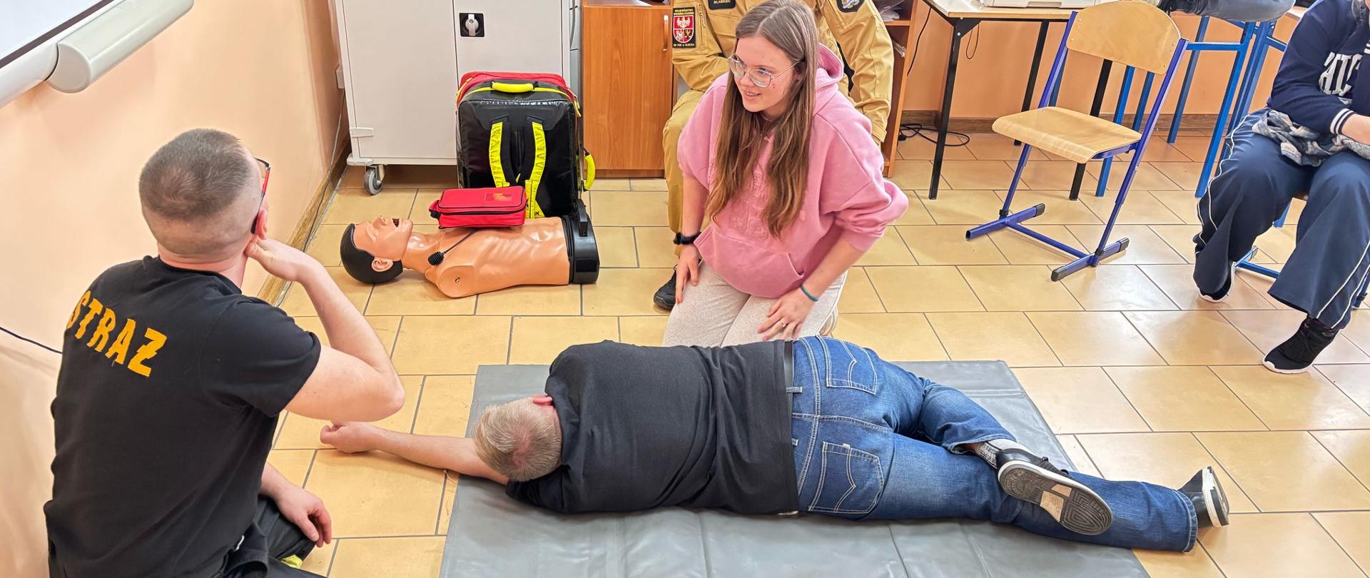 Na zdjęciu widoczny jest przebieg ze szkolenia Kwalifikowanej Pierwszej Pomocy w SP w Judzikach. Strażacy pokazują jak wygląda pozycja bezpieczna. Widoczny jest uczeń jako pozorant oraz uczennica jako osobą ćwicząca.