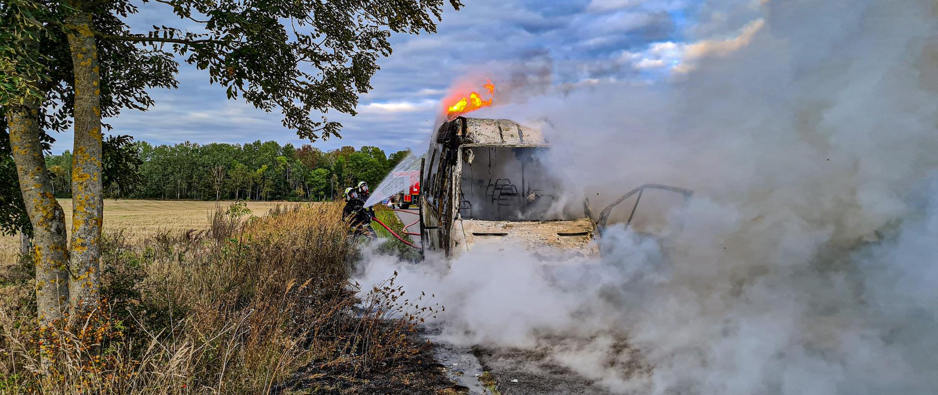Na fotografii w centrum w zadymieniu znajduje się palący się bus. Spośród dymu wydostają się jęzory ognia. Z tyłu busa po lewej stronie znajduje się dwóch strażaków, którzy podają prąd wody w natarciu na palącego się busa. Po lewej stronie fotografii znajduje się drzewo. W oddali znajduje się wóz strażacki oraz las. Zdjęcie wykonano w porze dziennej.