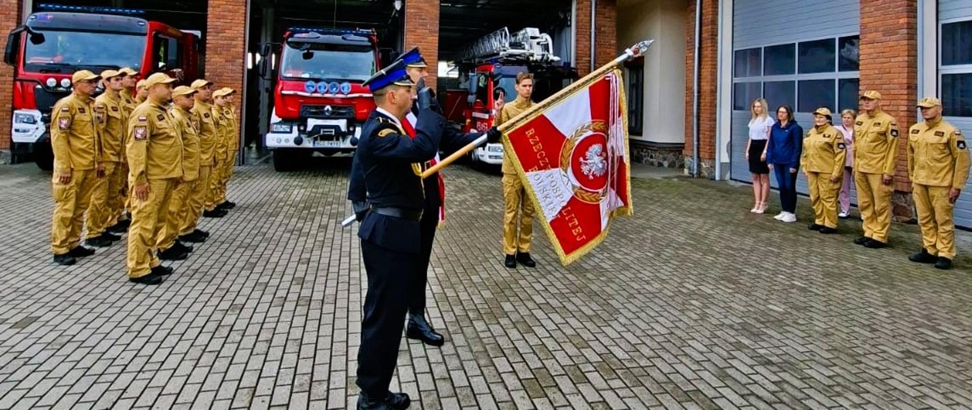 Zdjęcie przedstawia uroczysty moment składania ślubowania przez nowo przyjętego strażaka Macieja Bućkun do służby w Komendzie Powiatowej Państwowej Straży Pożarnej w Gołdapi oraz sztandar komendy. W tle można zaobserwować pozostałych strażaków biorących udział w uroczystej zbiórce oraz pojazdy pożarnicze zlokalizowane w garażu strażnicy