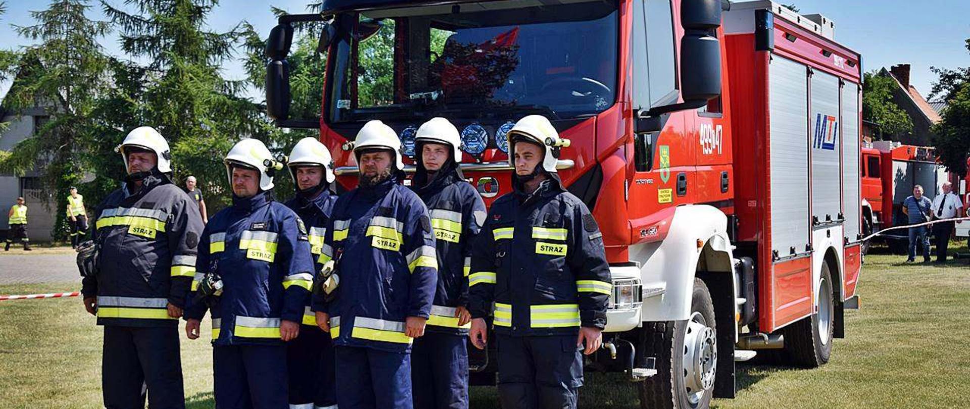 Na pierwszym planie stoją w dwuszeregu strażacy z OSP, ubrani w umundurowania bojowe - 6 osobowa załoga. Za nimi stoi wóz bojowy - średni gaśniczy, u góry z napisem OSP Sadki. Wszystko dzieje się na boisku, na murawie.