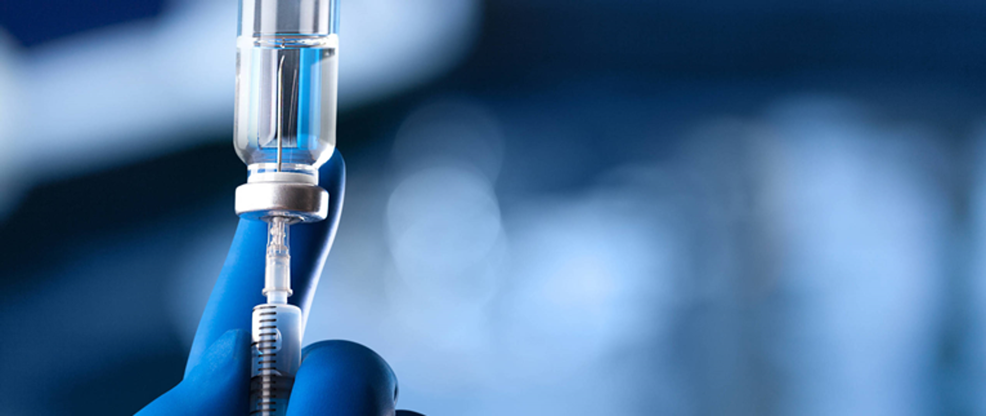 Fragment lewej dłoni w niebieskiej rękawiczce gumowej trzymająca fiolkę nabitą na igłę ze strzykawką. Wszystko na rozmytym niebieskim tle.