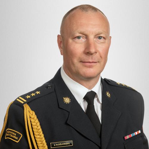 Portret mężczyzny w galowym mundurze Państwowej Straży Pożarnej, stojącego na jasnoszarym, neutralnym tle. Osoba jest ujęta od klatki piersiowej w górę, zwrócona lekko bokiem do aparatu i patrzy wprost przed siebie. Mundur ma złote elementy ozdobne, dystynkcje na naramiennikach, sznur galowy oraz odznaczenia na piersi.