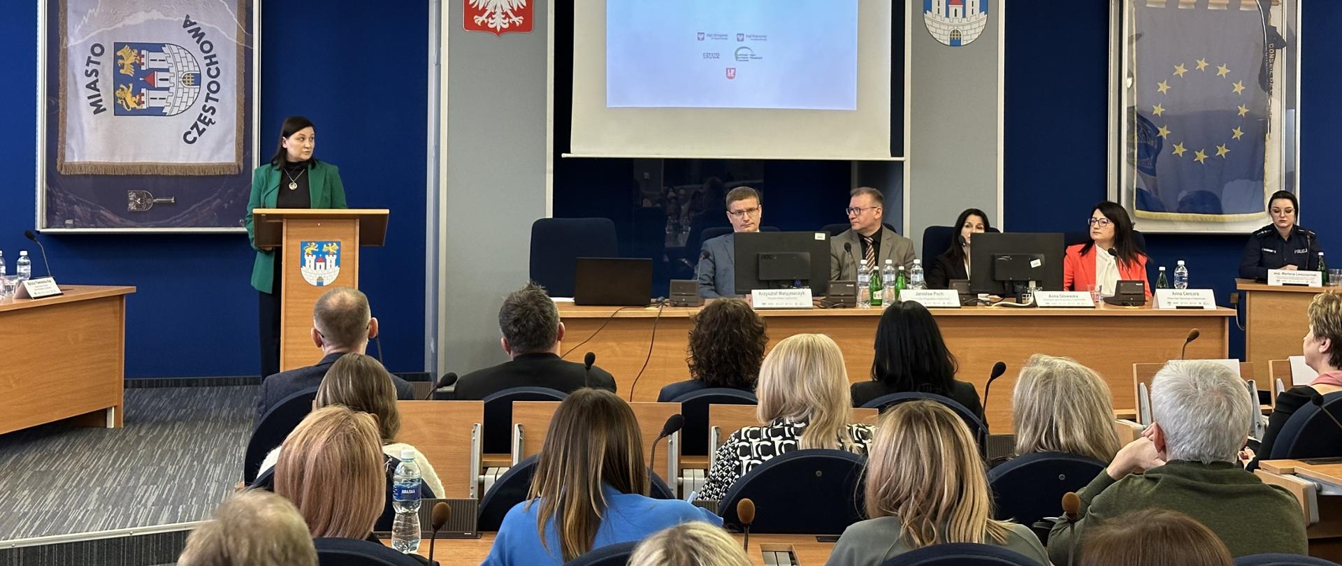 Obraz przedstawia ustawienie sali konferencyjnej z kilkoma osobami siedzącymi i jedną osobą stojącą na podium. Podium znajduje się z przodu sali, nieco po lewej stronie, i zajmuje je kobieta ubrana w zieloną kurtkę. Za nią na ścianie wywieszony jest sztandar z napisem "MIASTO CZĘSTOCHOWA" i herbem.
Na pierwszym planie znajduje się kilka rzędów uczestników siedzących przy biurkach, zwróconych twarzą do podium i ekranu projekcyjnego. Wśród uczestników przeważają kobiety, wśród których przeplata się kilku mężczyzn. Siedzą na krzesłach z zaokrąglonymi oparciami, a niektórzy mają przed sobą zeszyty lub papiery. Na jednym z biurek widoczna jest butelka wody.
Z przodu sali za długim biurkiem siedzi cztery osoby. Każdy z panelistów ma przed sobą mikrofon i tabliczkę z nazwiskiem. Wśród panelistów są dwaj mężczyźni i dwie kobiety. Nad panelistami na ekranie projekcyjnym wyświetlany jest slajd prezentacji.
