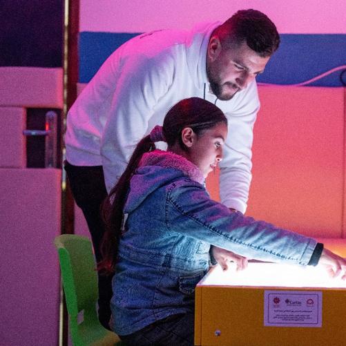 Kobieta w pomieszczeniu oświetlonym różową barwą siedzi przy stoliku, który ma świecący blat. Pokazuje coś. Nad nią pochyla się mężczyzna. 