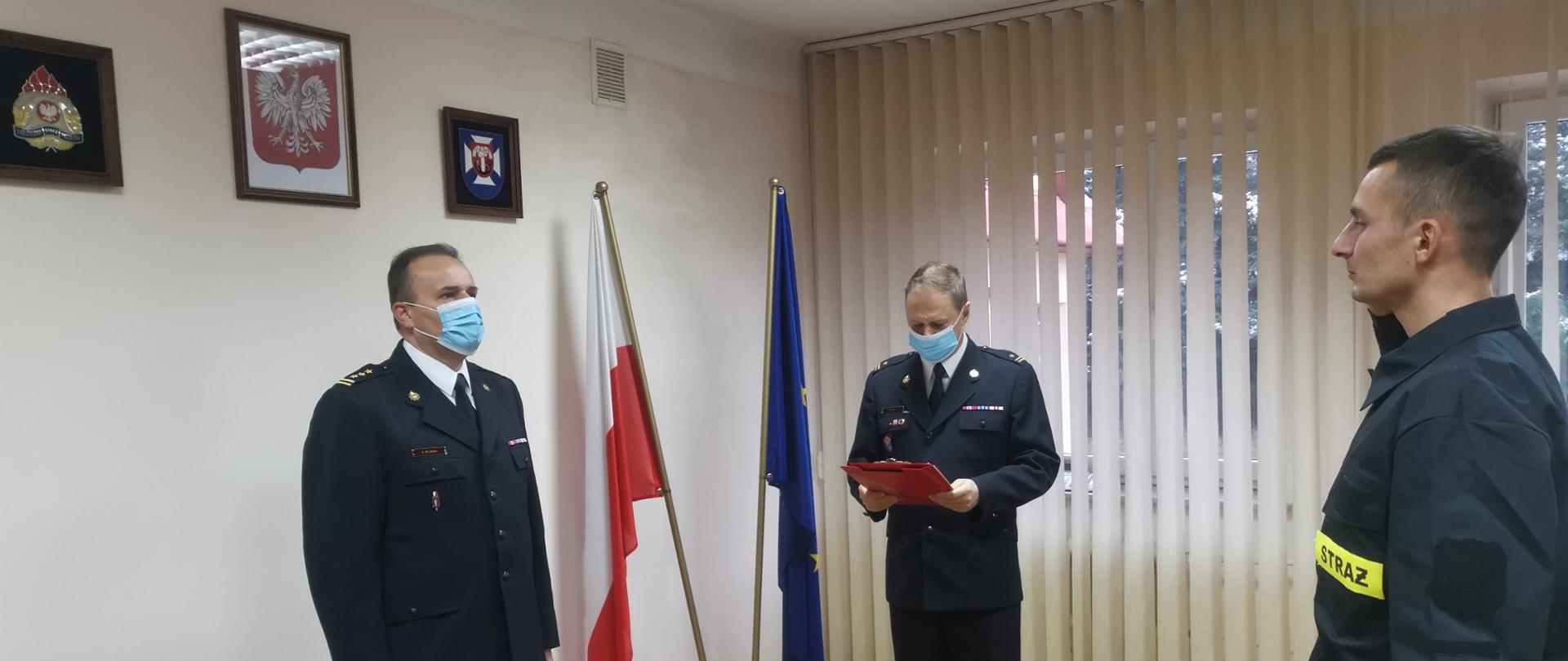 Zdjęcie przestawia pomieszczenie Komendy Powiatowej Państwowej Straży Pożarnej w Łańcucie Na ścianie godło Rzeczypospolitej Polskiej, herb powiatu łańcuckiego i logo Państwowej Straży Pożarnej. W pomieszczeniu znajdują się: starszy brygadier Jacek Rejman zastępca komendanta powiatowego Państwowej Straży Pożarnej w Łańcucie, młodszy brygadier Piotr Glac zastępca dowódcy Jednostki Ratowniczo-Gaśniczej w Łańcucie oraz nowo przyjęty strażak Rafał Reizer. Zastępca komendanta powiatowego i zastępca dowódcy JRG występują w umundurowaniu wyjściowym a strażak Rafał Reizer w umundurowaniu koszarowym. Strażak Rafał Reizer w postawie zasadniczej z podniesioną prawą ręką składa ślubowanie strażaka Państwowej Straży Pożarnej.