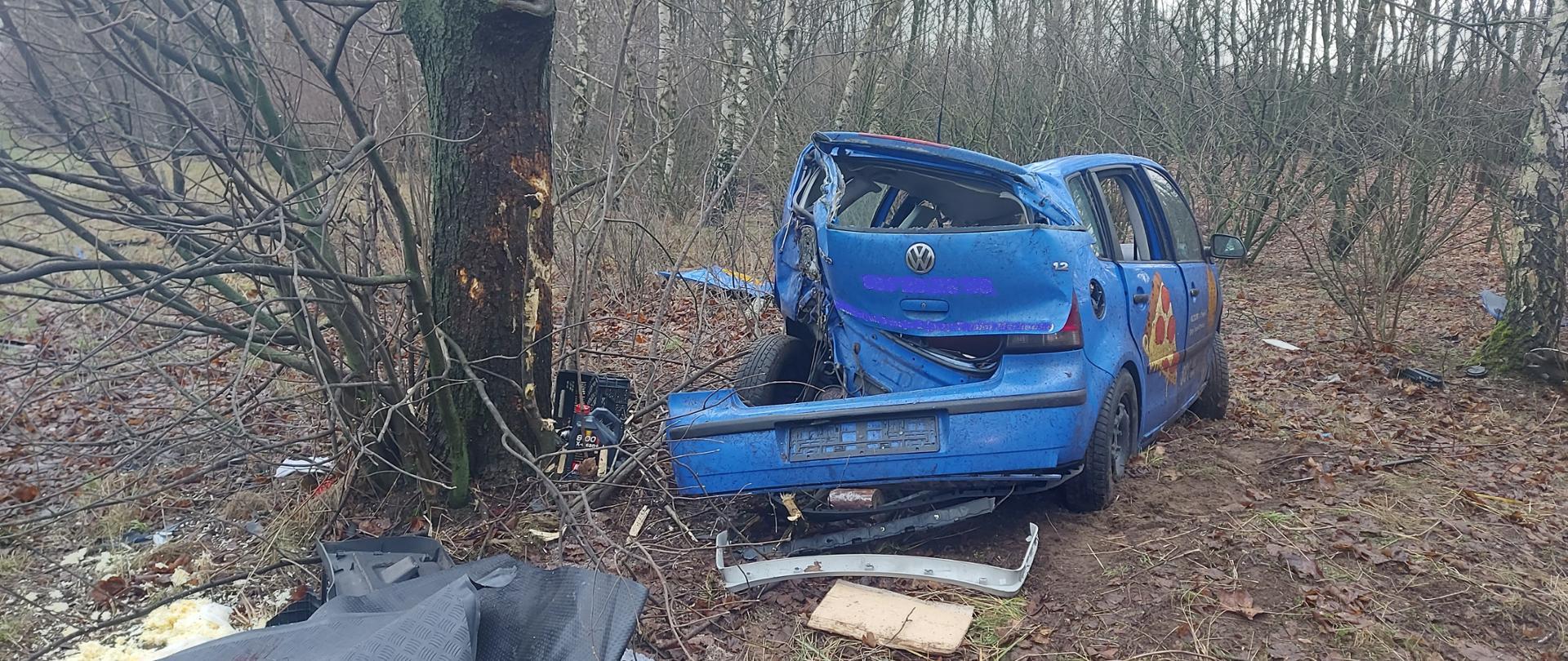 Zdjęcie przedstawia samochód osobowy koloru niebieskiego, który wcześniej uderzył w przydrożne drzewo. Samochód ma uszkodzony tył. Przed samochodem leżą części. 