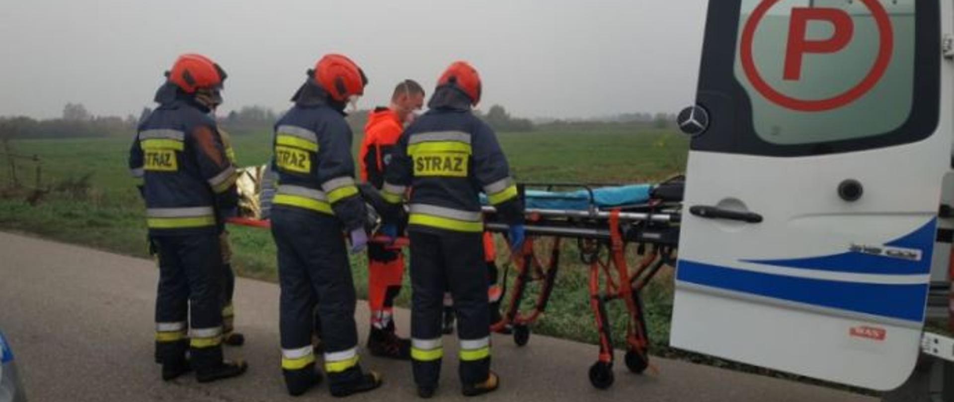 Na zdjęciu strażacy pomagają ratownikom medycznym włożyć nosze z osobą poszkodowaną do karetki pogotowia.