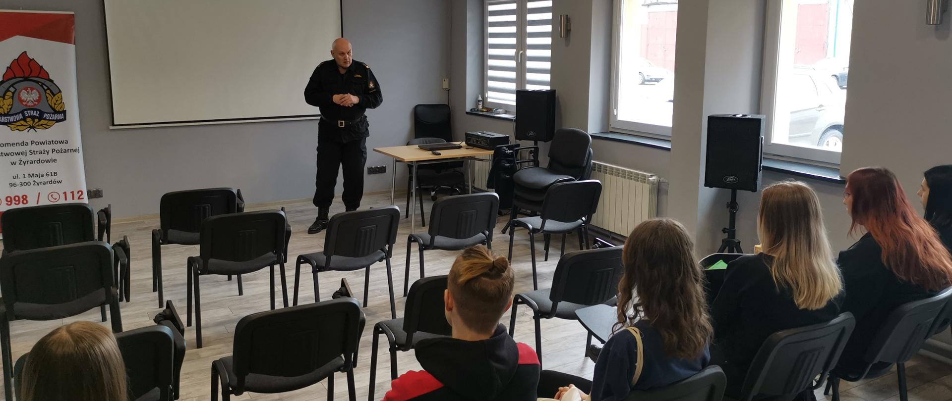 Na sali wykładowej stoi funkcjonariusz. Przed nim na krzesłach siedzi pięć dziewczyn i jeden chłopak, w tle widać ekran rzutnika.