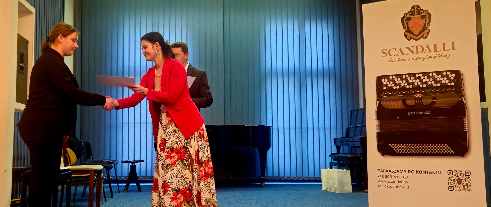 Na zdjęciu widoczne są dwie kobiety stojące na scenie. Kobieta po lewej, ubrana w ciemny strój, podaje rękę kobiecie po prawej, ubranej w czerwoną marynarkę i długą spódnicę w kwiaty, która wręcza jej dokument. W tle, za nimi, stoi mężczyzna. Po prawej stronie sceny widoczny jest pionowy baner z logo „SCANDALLI” i wizerunkiem akordeonu. Scena ma niebieską wykładzinę i niebieskie żaluzje w tle.