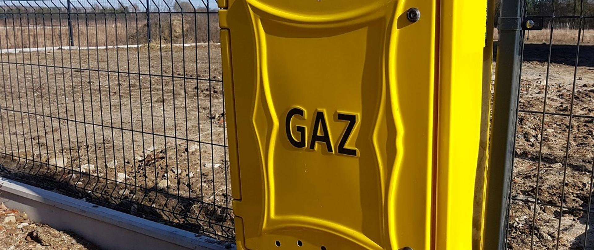 Zdjęcie przedstawia skrzynkę gazową w kolorze żółtym zamocowaną w panelu ogrodzeniowym, na skrzynce napis gaz w kolorze czarnym