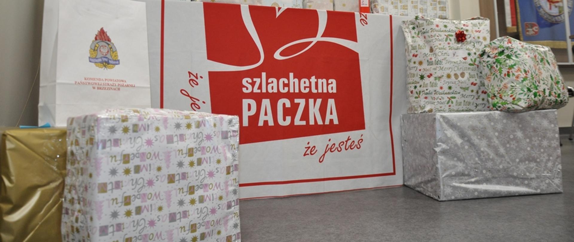 Zakupione prezenty przez brzezińskich strażaków PSP ok. 20 pakunków zapakowanych w świąteczny papier. Duży plakat z napisem "Szlachetna Paczka". Prezenty w pomieszczeniu komendy PSP w Brzezinach.