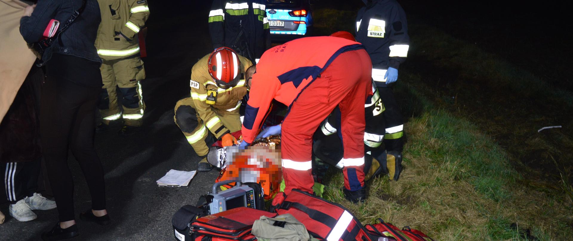 Wypadek w Stwolnie. Na jezdni - na desce ortopedycznej leży ranna kobieta, wcześniej zaopatrzona przez strażaków. Nad nią pochyla się lekarz Lotniczego Pogotowia Ratunkowego, który przeprowadza badanie. Po obu stronach poszkodowanej klęczą strażacy pomagający medykowi. Dwaj inni strażacy stoją z tyłu poszkodowanej. W tle widoczny radiowóz policyjny z włączonymi niebieskimi światłami błyskowymi. Z lewej strony stoi kobieta odwrócona plecami, obok niej strażak.