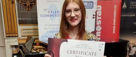na pierwszym planie rudowłosa dziewczyna w okularach trzymająca w rękach dyplom na którym po lewej stronie wiolonczela pośrodku napis z gratulacjami za III miejsce wiolonczelowym
