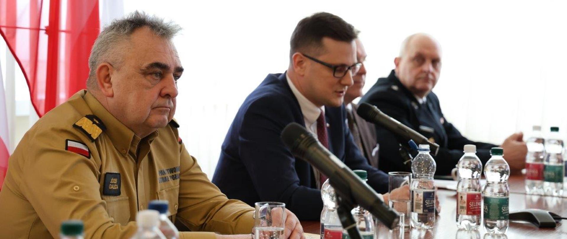 Na pierwszym planie siedzi mężczyzna w piaskowym mundurze PSP z plakietką imienną „Józef Galica”, mający skupiony i poważny wyraz twarzy. Obok niego znajduje się mężczyzna w garniturze i okularach, który mówi do mikrofonu. W tle siedzą inni uczestnicy spotkania, w tym osoba w mundurze strażackim. Na stole ustawione są mikrofony, butelki wody oraz dokumenty. W tle widoczna jest polska flaga, co podkreśla oficjalny charakter wydarzenia. Atmosfera spotkania sugeruje ważne rozmowy dotyczące działań straży pożarnej lub współpracy między instytucjami.