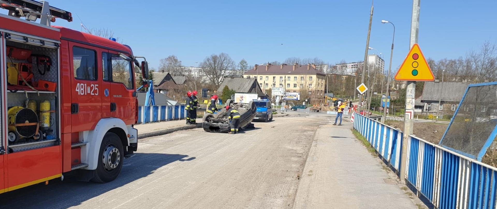 Na pierwszym planie widzimy samochód straży pożarnej, który zabezpiecza miejsce zdarzenia. W tle zdjęcia znajduje się samochód osobowy BMW, który dachował. Za nim widzimy patrol Policji. Wokół uszkodzonego pojazdu znajdują się strażacy, którzy rozłączają zasilanie akumulatorowe.