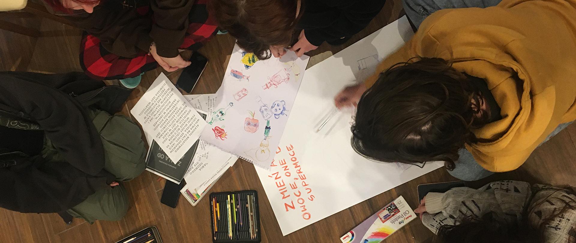 Na zdjęciu uczniowie pochylających się tworząc pracę plastyczną na brystolu prezentującą wybrane zagadnienia z warsztatów psychologicznych. Ujęcie wykonane z góry, widać tylko czubki głów uczniów.