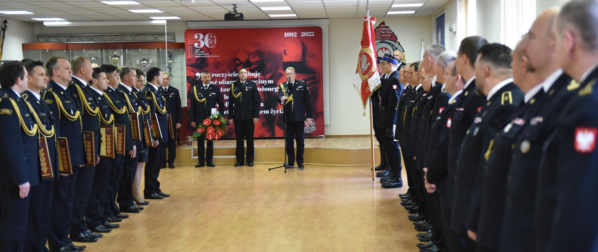 strażacy stojący na zbiórce w sali wszyscy w mundurach wyjściowych, stoją w 2 grupach po lewej i prawej, trzech stoi na środkowym planie za nimi baner 30-lecia państwowej straży pożarnej
