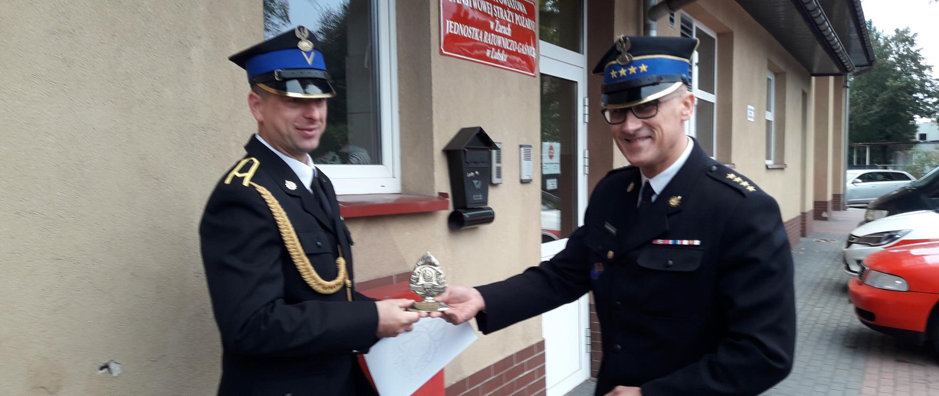 Na pierwszym planie widoczny jest Komendant Powiatowy wręczający statuetkę dla strażaka, w tle widoczny jest budynek jednostki w Lubsku.