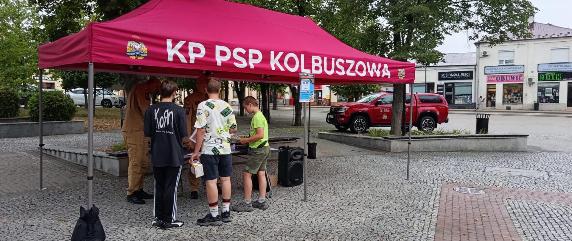 Rynek Kolbuszowski. Na środku stoi czerwony namiot typu wezyr z odsłoniętymi bokami. Na namiocie widnieje napis KP PSP Kolbuszowa. Pod namiotem znajdują się stół a za stołem stoją dwaj strażacy w piaskowych ubraniach służbowych. Przed stołem stoi trzech chłopaków w ubraniach cywilnych w wieku od 13 do 17 lat. Funkcjonariusze przedstawiają materiały z warsztatów. Na stole znajduję się laptop a zboku stołu stoi kolumna głośnikowa. Parę metrów za namiotem na placu rynku stoi strażackie czerwone terenowe służbowe auto. Plac rynku jest wybrukowany a dodatkowo gdzieniegdzie rosną duże drzewa. Za namiotem widać też że częściowo plac jest pokryty trawnikiem i małymi krzewami na murkach. W tle zdjęcia widać ściany kamienic rynku w których znajdują się sklepów z widocznymi szyldami. 
