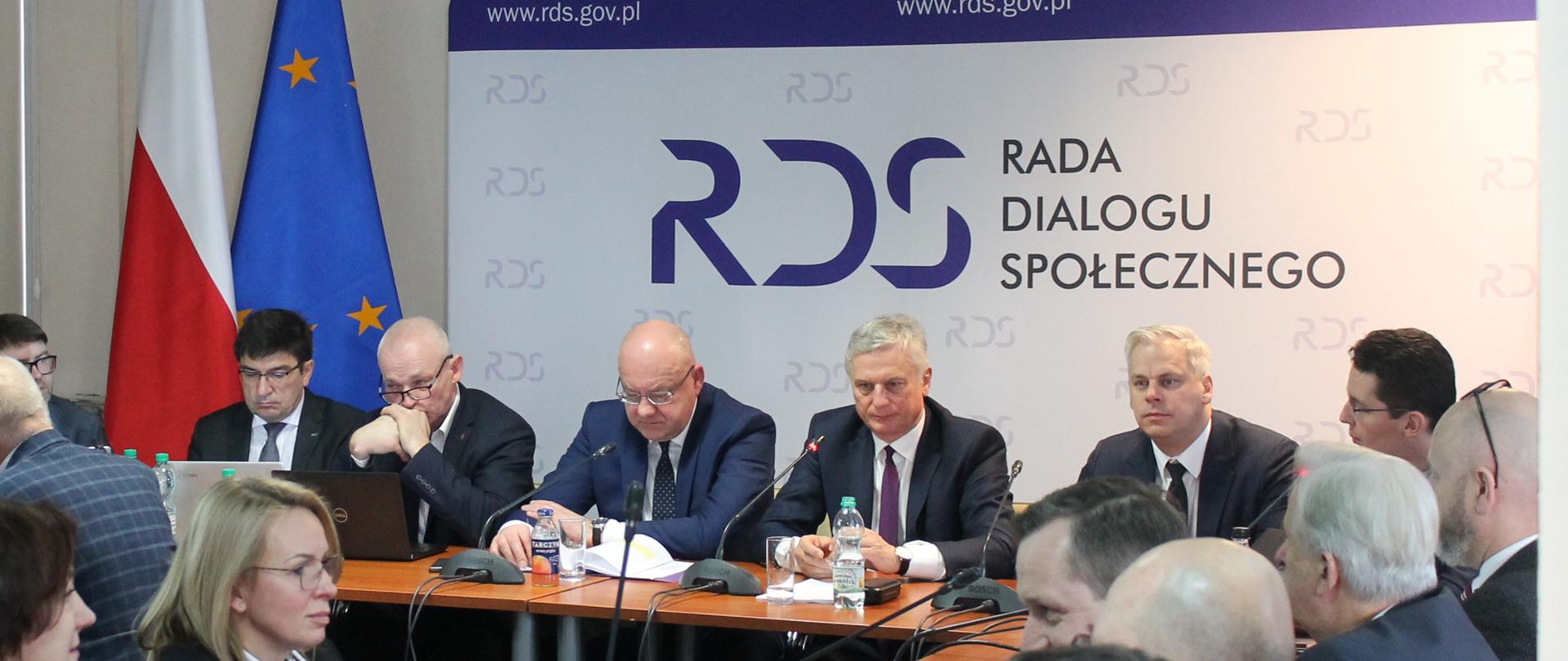 Zdjęcie przedstawia stół prezydialny za którym siedzą: Pan Karol Rabenda, Podsekretarz Stanu w Ministerstwie Aktywów Państwowych, Współprzewodniczący Zespołów ze strony rządowej; Pan Ryszard Wasiłek, Współprzewodniczący Zespołu Trójstronnego ds. Branży Energetycznej ze strony pracodawców; Pan Wojciech Ilnicki, Współprzewodniczący Zespołu Trójstronnego ds. Branży Węgla Brunatnego ze strony związków zawodowych oraz Pan Sławomir Wochna, Współprzewodniczący Zespołu Trójstronnego ds. Branży Węgla Brunatnego ze strony pracodawców. W tle siedzących znajduje się baner z napisem Rada Dialogu Społecznego. Po lewej stronie zdjęcia stoją dwie flagi: Polska i UE. Nad banerem widnieje Godło Polski.