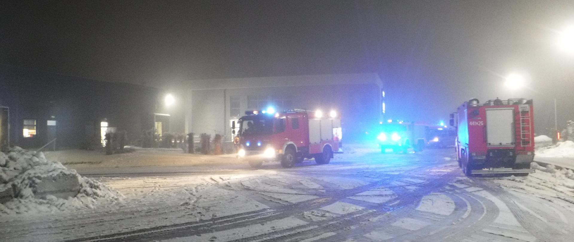 Na zdjęciu widać samochody straży pożarnej na tle magazynu, w którym doszło do pożaru. Jest ciemno a na ziemi leży śnieg.
