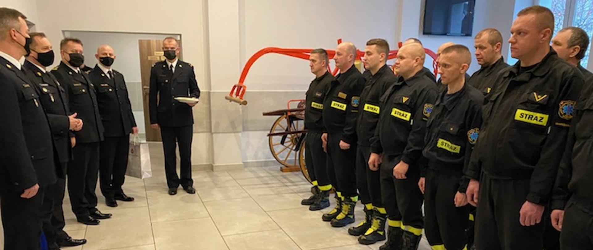 Po lewej kadra kierownicza Państwowej Straży Pożarnej , po prawej zmiana służbowa na spotkaniu przedświątecznym W tle zabytkowa sikawka