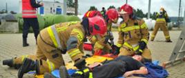 Strażacy udzielający pomocy medycznej pozorowanej osobie poszkodowanej. W tle widoczni są inni strażacy biorący udział w ćwiczeniach.