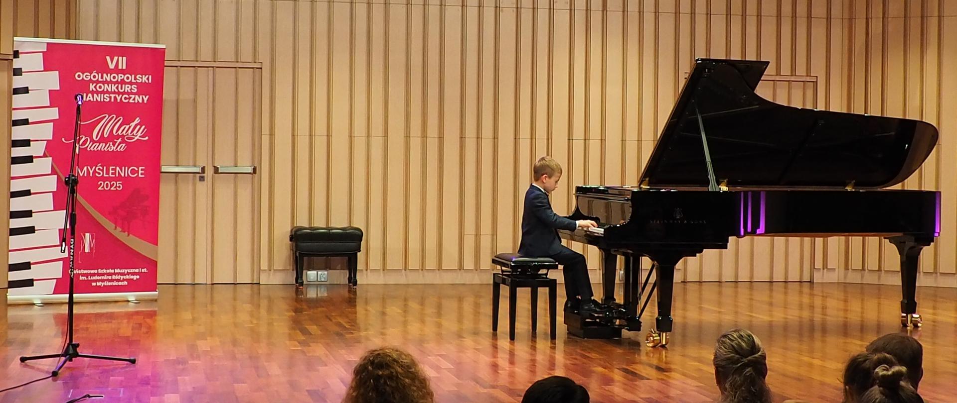 Uczeń gra na fortepianie na scenie w sali koncertowej, obok plakat z tekstem: VII Ogólnopolski Konkurs Pianistyczny „Mały Pianista” Myślenice 28-29 maja 2025 Państwowa Szkoła Muzyczna I stopnia im. Ludomira Różyckiego w Myślenicach 