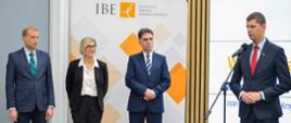 Wiceminister Piontkowski stoi i mówi do mikrofonu, obok niego stoi dwóch mężczyzn w garniturach i ubrana na czarno kobieta, za nimi ścianka z napisem IBE.